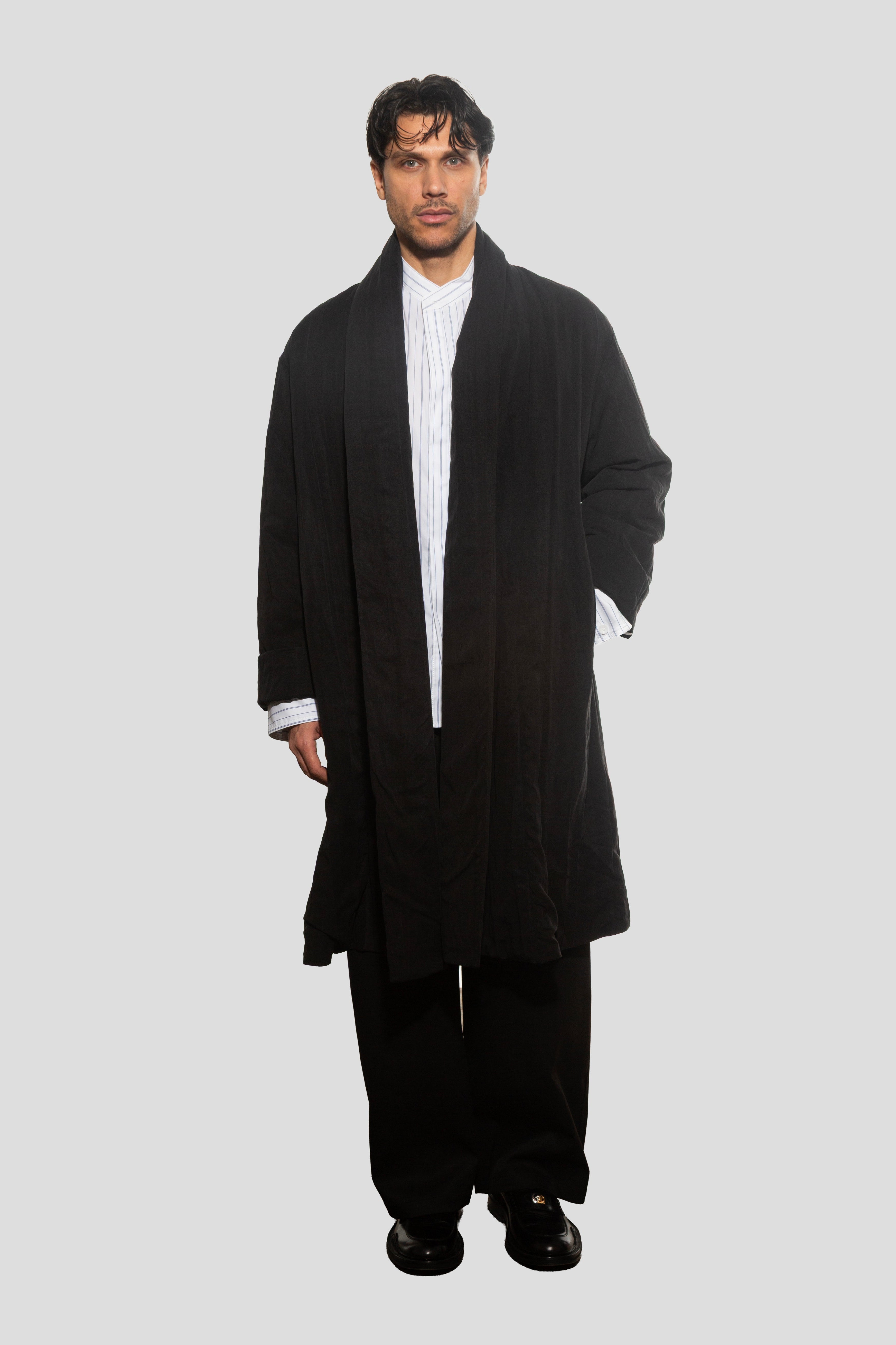 Man wearing a long black coat over a white shirt on a light gray background
